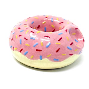 May include: A ceramic donut-shaped object with pink icing and colourful sprinkles. The bottom of the donut is a light yellow colour. The sprinkles are red, blue, and yellow. The donut is on a white background.