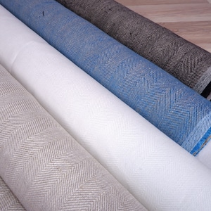 Op de afbeelding: Gerolde stoffen rollen in diverse kleuren, waaronder wit, blauw en grijs, met een visgraatpatroon. De stof lijkt van een gemiddeld gewicht te zijn, geschikt voor bekleding of kleding.
