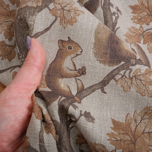 Op de afbeelding: Een beige linnen stof met een bruine eekhoorn en bladeren erop gedrukt. De stof is getextureerd en heeft een natuurlijke uitstraling.