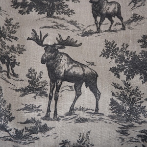 May include: A fabric with a black and white toile pattern featuring moose and forest scenes. The moose have large antlers and are set against a backdrop of trees and hills. The fabric has a neutral, beige background.