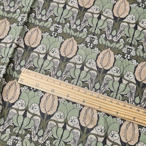 May include: Close-up of fabric with a repeating pattern of owls, leaves, and berries in shades of green, beige, and white. A wooden ruler with centimetre markings is placed diagonally across the fabric, measuring up to 19 cm.