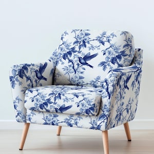Op de afbeelding: Witte fauteuil met een blauw bloemen- en vogelpatroon. De stoel heeft een afgeronde rugleuning, armleuningen en lichtgekleurde houten poten. De stof heeft een gedetailleerd ontwerp van bloemen en vogels.
