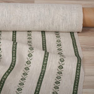 Può includere: Un rotolo di tessuto di lino naturale con strisce verticali. Le strisce alternano tra bande verde scuro piene e bande con un motivo floreale e geometrico in verde scuro. Il tessuto è parzialmente srotolato, rivelando la trama e il motivo.