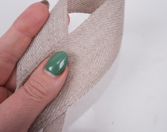 May include: A close-up of a beige linen ribbon, folded in half, with a hand holding the ribbon. The hand has green nail polish.