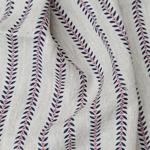 Può includere: Primo piano di un tessuto beige a righe verticali. Ogni striscia presenta un motivo ripetuto di foglie blu scuro e piccoli accenti rossi. Il tessuto ha un aspetto strutturato, che suggerisce una fibra naturale come il lino o il cotone.