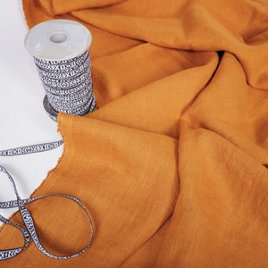 May include: A spool of black and white patterned trim sits on a folded piece of mustard yellow linen fabric.