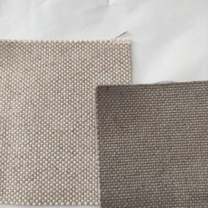 Könnte beinhalten: Zwei Stoffmuster in neutralen Tönen. Eines ist hellbeige mit engem Gewebe, das andere ist ein dunkleres Graubraun mit ähnlichem Gewebe. Die Texturen sind sichtbar, was auf Polster- oder Vorhangmaterial hindeutet.