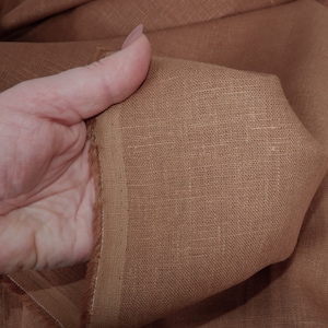 May include: Close-up of a brown linen fabric with a slightly textured surface. The fabric is folded and held in a hand.