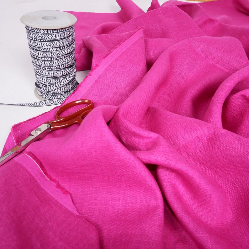 May include: A close-up of a bright pink linen fabric with a pair of scissors and a spool of black and white patterned ribbon.
