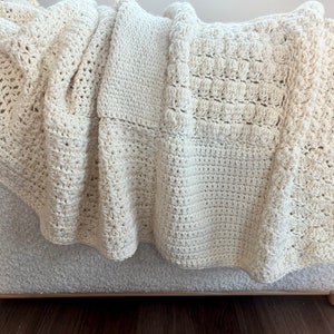 May include: A cream-coloured crocheted throw blanket with different stitch patterns. The blanket is draped over a sofa.