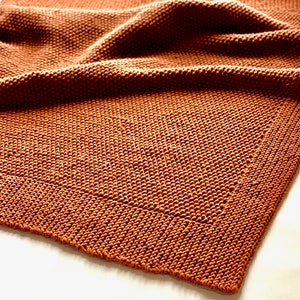 May include: A brown knitted throw blanket with a textured weave. The blanket is folded and draped over a white surface.