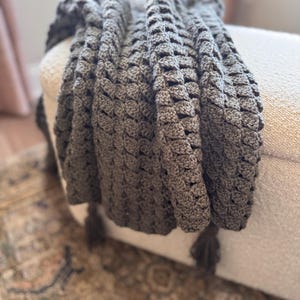 Può includere: Una coperta grigia all'uncinetto, con texture e nappe. La coperta è drappeggiata su un pouf bianco, con un tappeto fantasia visibile in primo piano. Il motivo all'uncinetto crea un effetto dimensionale in rilievo.
