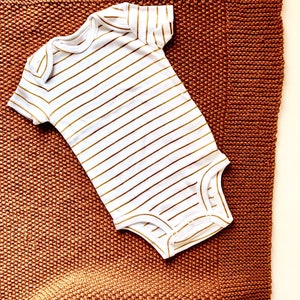 May include: A white baby onesie with gold stripes on a brown knitted blanket.