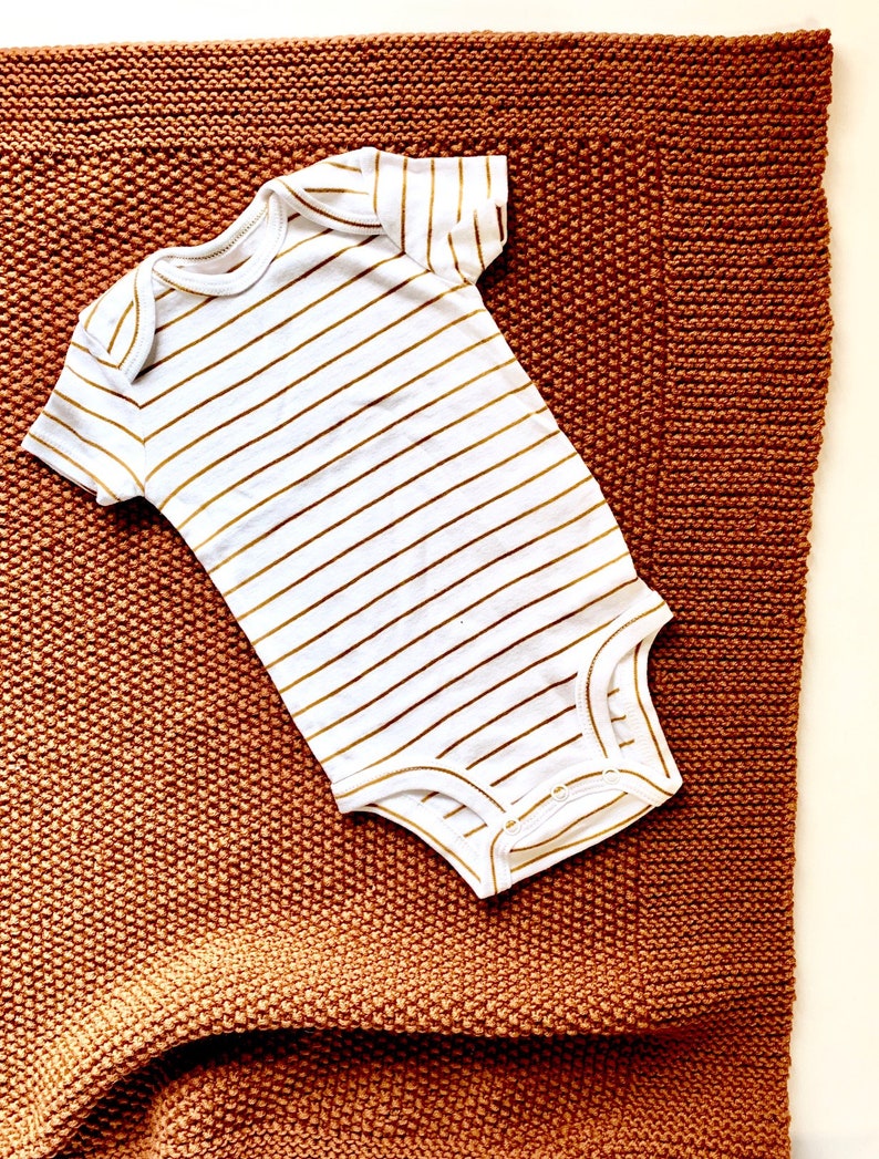 May include: A white baby onesie with gold stripes on a brown knitted blanket.