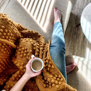 May include: A person relaxing on a sofa with a mustard yellow crocheted throw blanket and a white mug of coffee. The person is wearing blue jeans and has their feet up on the sofa.