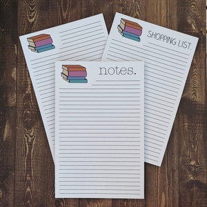 May include: Three white lined notepads with a stack of colorful books illustration. The top notepad says "SHOPPING LIST." The middle notepad says "notes."