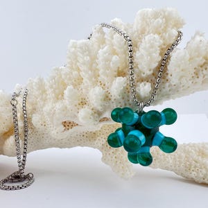 May include: A silver chain necklace with a pendant featuring a teal and blue molecular design. The necklace is displayed on a piece of white coral against a white background. A second silver chain is also visible.