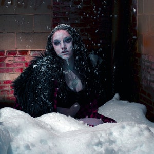 May include: A woman with dark hair and pale skin is wearing a black fur coat and a red dress. She is sitting in a snowy alleyway with brick walls. Snow is falling around her.