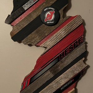 May include: A wooden outline of the state of New Jersey decorated with hockey sticks and a New Jersey Devils hockey puck. The puck has the team logo and the words "New Jersey Devils".