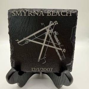 May include: A square black slate coaster featuring an engraved airport runway diagram for Smyrna Beach. The text Smyrna Beach is etched at the top, and the date 12/1/2007 is engraved at the bottom. The coaster has a rough, natural edge texture.