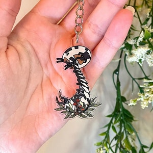 May include: A black and white cartoon scorpion keychain with red accents and green leaves. The scorpion is curled up in a defensive position.