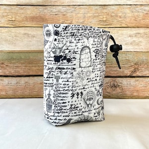 May include: A white drawstring bag with a black and white print of various symbols, including a hand, an eye, and a sun. The bag has a black drawstring.