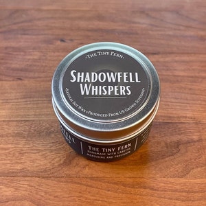 May include: A silver tin candle with a black label that reads "THE TINY FERN. SHADOWFELL WHISPERS. NATURAL SOY WAX. PRODUCED FROM US GROWN SOYBEANS."