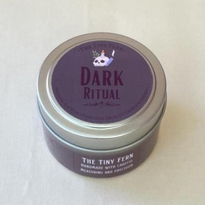 May include: A silver tin candle with a purple label that reads "The Tiny Fern" and "Dark Ritual". The label also features a skull with a candle flame on top.