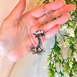 May include: A black and white cartoon mushroom keychain with a silver chain. The mushroom has a white and black spotted cap and a long, thin stem with a brown base. The mushroom is surrounded by green leaves and red flowers.