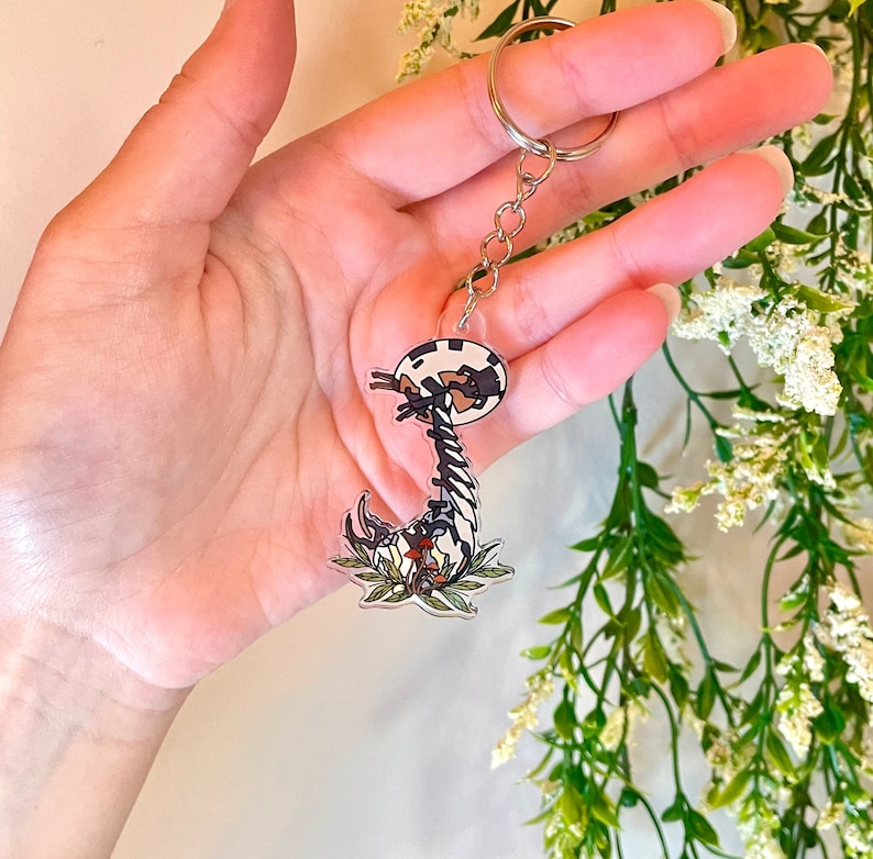 May include: A black and white cartoon mushroom keychain with a silver chain. The mushroom has a white and black spotted cap and a long, thin stem with a brown base. The mushroom is surrounded by green leaves and red flowers.