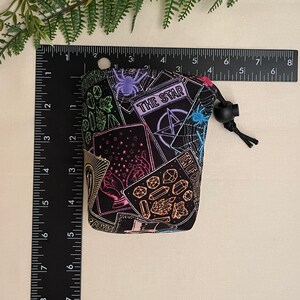 Op de afbeelding: Een zwarte trekkoordtas met een kleurrijk tarotkaartontwerp. De tas heeft verschillende tarotkaartsymbolen, waaronder de kaart "THE STAR", een pentagram en een spinnenweb. De tas is ongeveer 10 cm hoog.