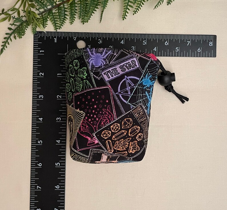 Op de afbeelding: Een zwarte trekkoordtas met een kleurrijk tarotkaartontwerp. De tas heeft verschillende tarotkaartsymbolen, waaronder de kaart "THE STAR", een pentagram en een spinnenweb. De tas is ongeveer 10 cm hoog.