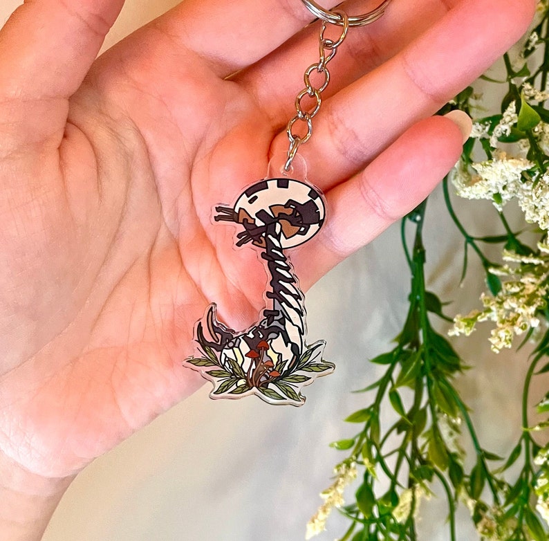 May include: A clear acrylic keychain featuring a black and white illustration of a mushroom creature with a long, segmented body. The creature is surrounded by green leaves and red mushrooms.