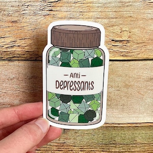 May include: A sticker featuring a jar of "Anti-Depressants." The jar is filled with green, geometric shapes. The label on the jar reads "Anti-Depressants" in a bold, sans-serif font. The jar has a brown cap and is set against a wooden background.