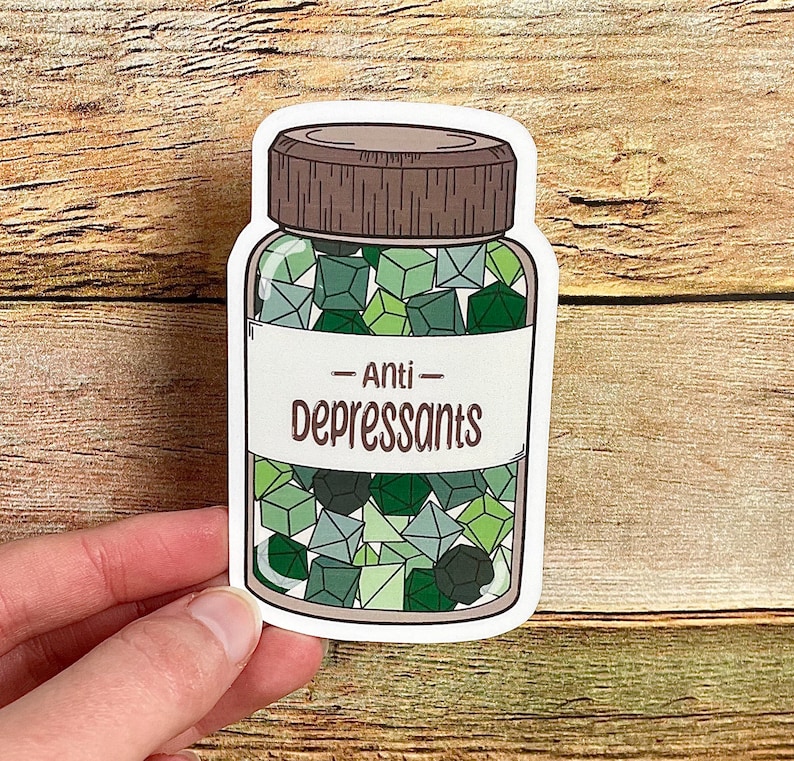 May include: A sticker featuring a jar of "Anti-Depressants." The jar is filled with green, geometric shapes. The label on the jar reads "Anti-Depressants" in a bold, sans-serif font. The jar has a brown cap and is set against a wooden background.