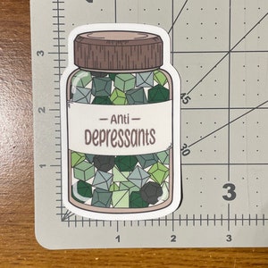 May include: A sticker featuring a jar of dice labeled "Anti-Depressants." The jar is filled with various green and gray dice. The jar has a brown lid and a white label with the text in a serif font.