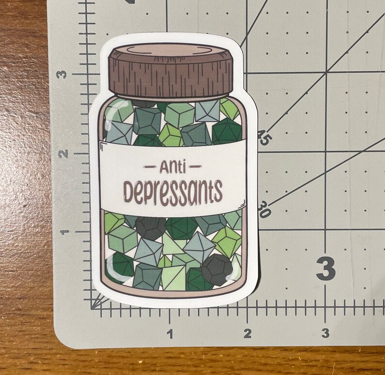 May include: A sticker featuring a jar of dice labeled "Anti-Depressants." The jar is filled with various green and gray dice. The jar has a brown lid and a white label with the text in a serif font.