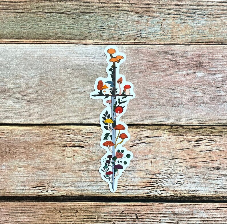 May include: A sticker featuring a sword design with colorful mushrooms and foliage. The sword has a silver blade and a black hilt. The mushrooms are in shades of orange, red, yellow, and brown. The sticker is on a wooden surface.