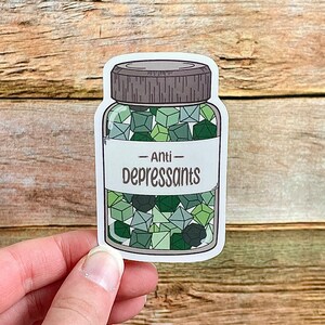 May include: A sticker featuring a jar of "Anti-Depressants." The jar is filled with green geometric shapes. The label on the jar reads "Anti-Depressants" in a bold, sans-serif font. The jar has a gray lid and is outlined in black.
