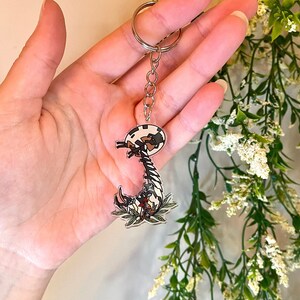 May include: A clear acrylic keychain featuring a black and white cartoon centipede with red and white flowers. The keychain is attached to a silver chain.