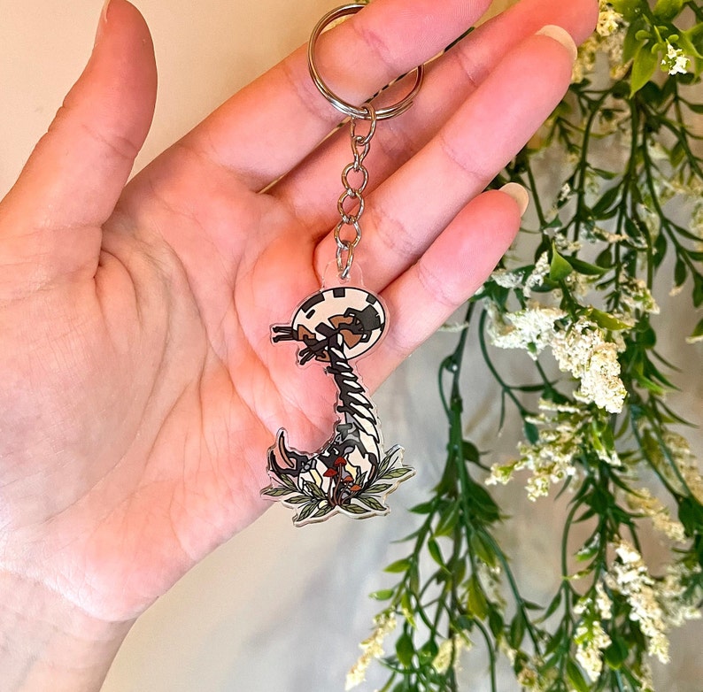 May include: A clear acrylic keychain featuring a black and white cartoon centipede with red and white flowers. The keychain is attached to a silver chain.