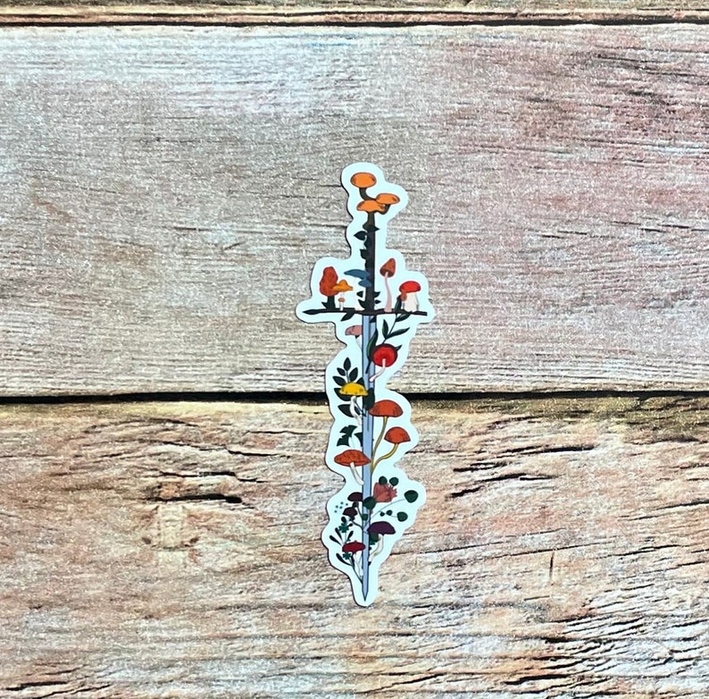 May include: A sticker featuring a sword design with colorful mushrooms and foliage. The sword has a dark gray blade and a black hilt. The mushrooms are various colors, including orange, red, and yellow. The sticker is on a wooden surface.