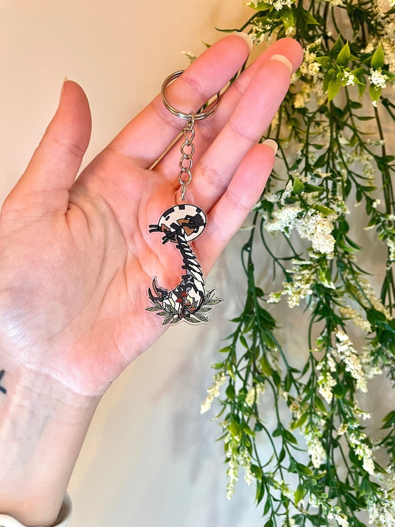 May include: A black and white cartoon scorpion keychain with red accents and green leaves. The scorpion is curled up in a defensive position.