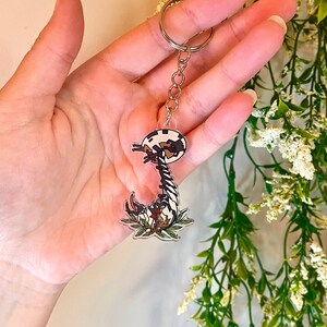 May include: A black and white cartoon keychain featuring a creature with a long neck and a round head. The keychain is attached to a silver key ring and has a green and brown leafy design around the bottom.