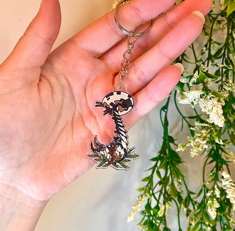 May include: A black and white cartoon keychain featuring a creature with a long neck and a round head. The keychain is attached to a silver key ring and has a green and brown leafy design around the bottom.