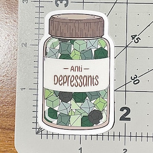 May include: A sticker featuring a clear jar with a brown lid, filled with green and gray polyhedral dice. The label reads "Anti-Depressants" in a brown, handwritten-style font. The sticker is on a white background.