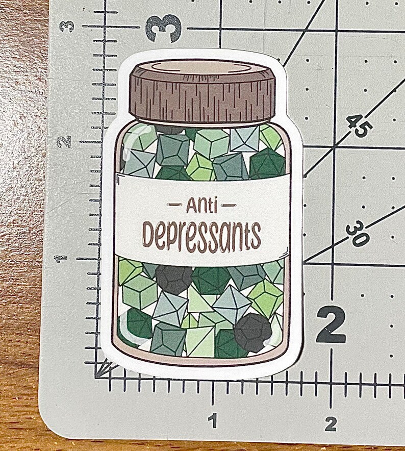 May include: A sticker featuring a clear jar with a brown lid, filled with green and gray polyhedral dice. The label reads "Anti-Depressants" in a brown, handwritten-style font. The sticker is on a white background.