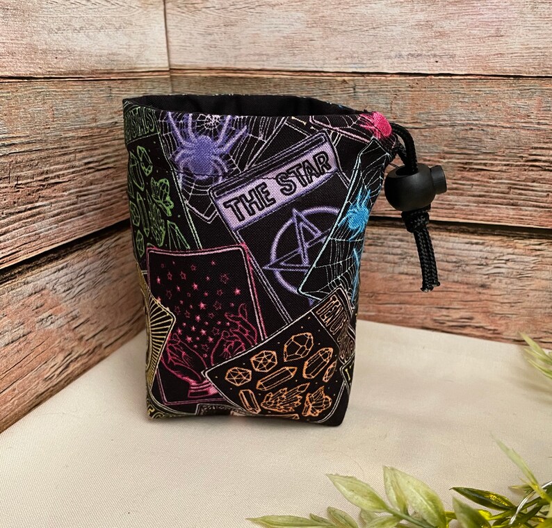 Op de afbeelding: Zwarte stoffen dobbelsteen tas met een trekkoord. De tas is bedrukt met een kleurrijk ontwerp van tarotkaarten, kristallen en een pentagram. De tekst "The Star" is zichtbaar op een van de kaarten.