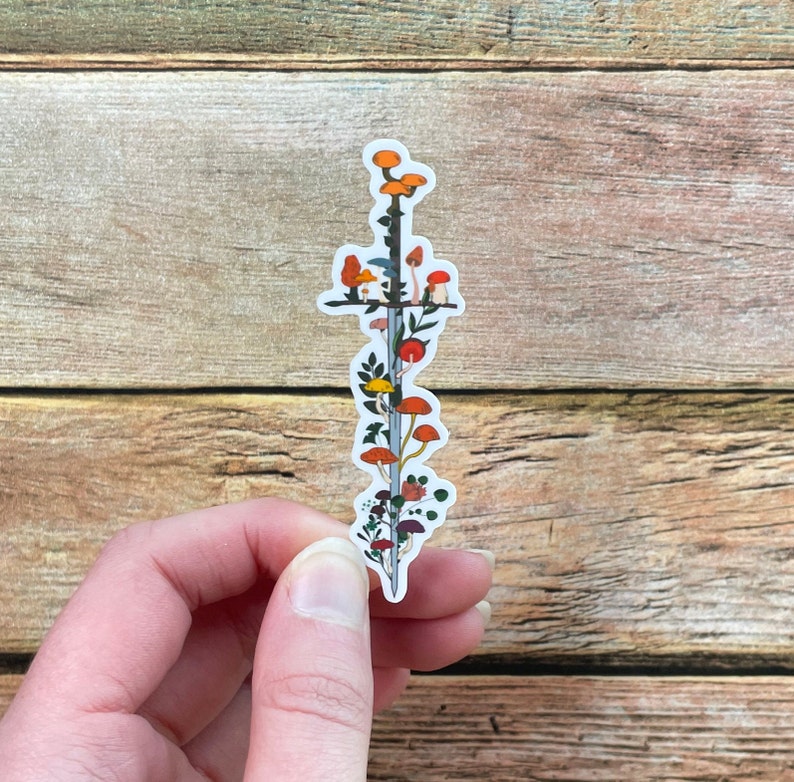 May include: A sticker featuring a sword design with colorful mushrooms and foliage. The sticker has a white background and is held by a hand. The mushrooms are orange, red, and brown, with green and yellow leaves.