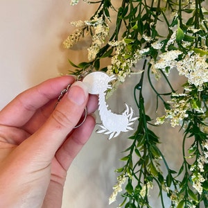May include: A white, detailed, floral design on a silver key chain. The design is of a scorpion with its tail curled up.